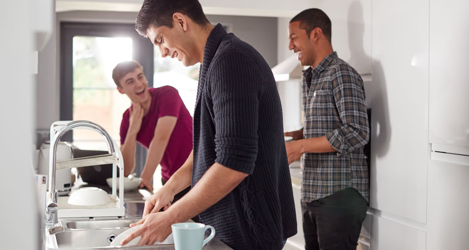 Group Of Male College Students In Shared House Kitchen Washing Up And Hanging Out Together