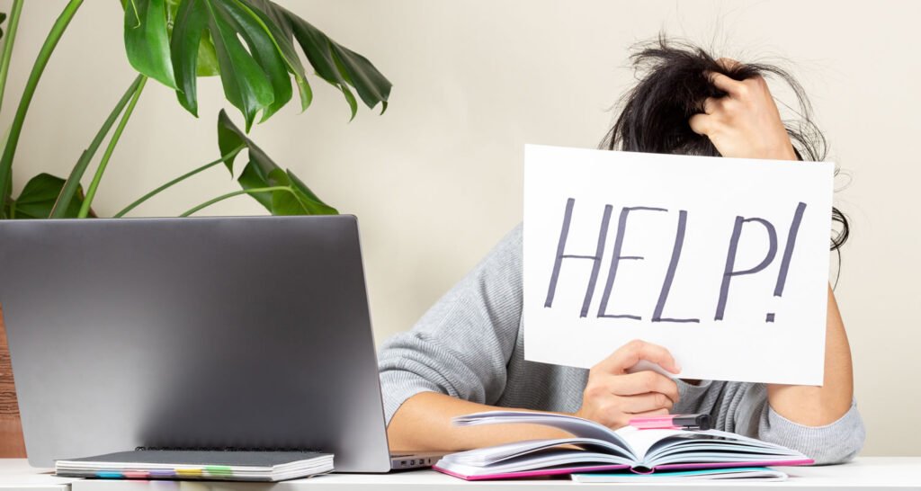 Tired frustrated student sitting at table and holding card with word Help.